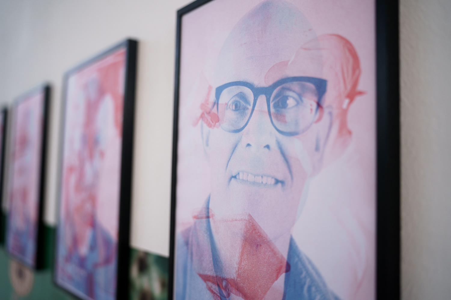 Close-up of a framed risograph poster with a red and blue duotone portrait of a man wearing glasses, part of a row of posters on the wall