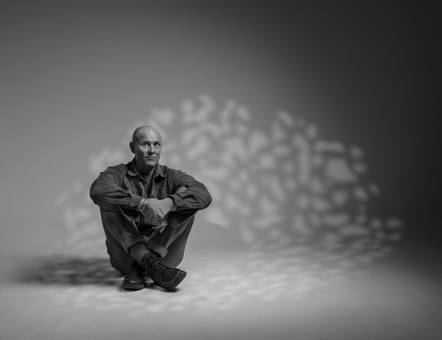 Black-and-white portrait of a man sitting on the floor hugging his knees, with dappled shadow patterns cast on the wall behind him