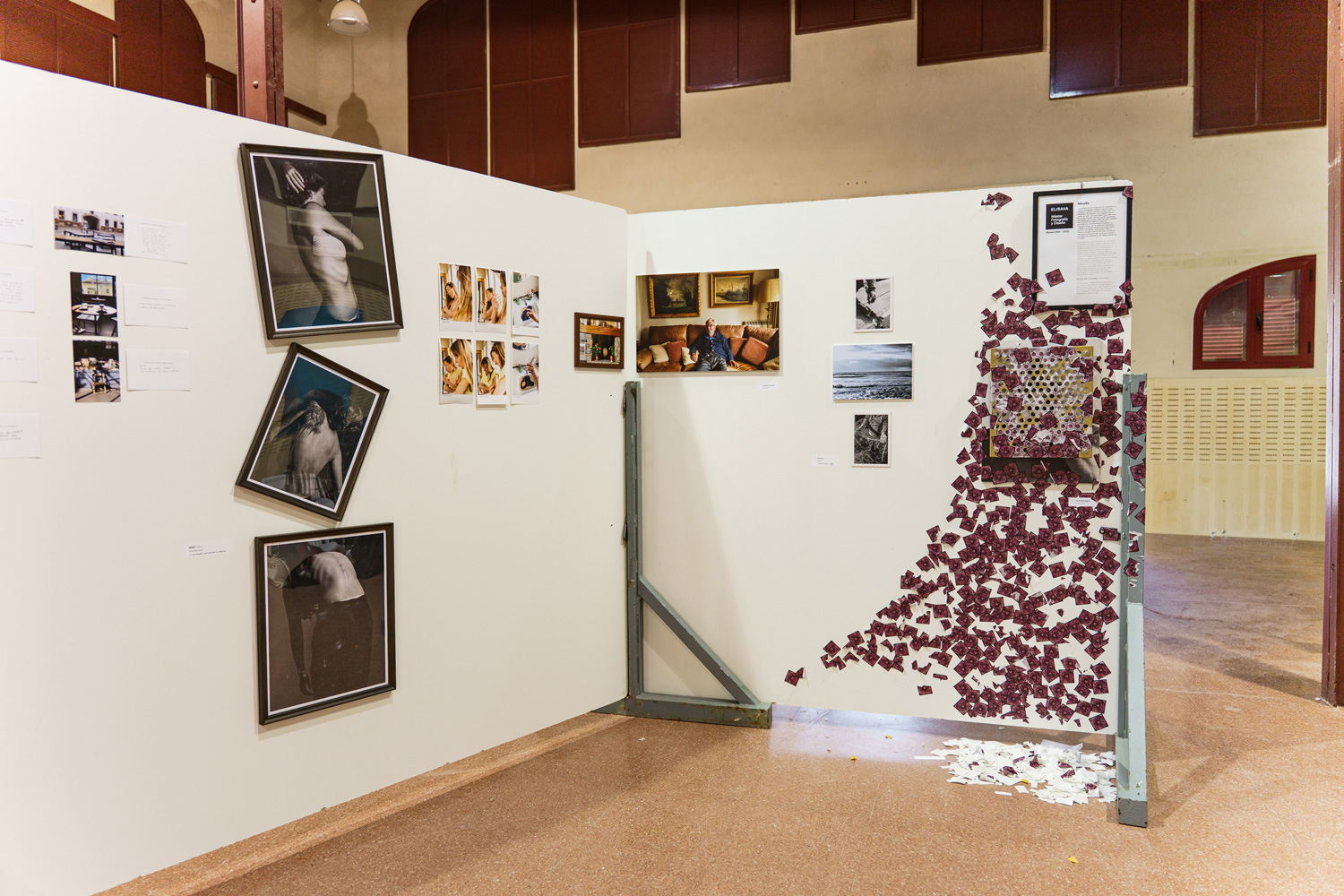 Wide view of the gallery wall for Overstimulated, showing a perforated gold panel covered in scattered pink QR code stickers spilling down the white wall beside framed black-and-white and color photographs